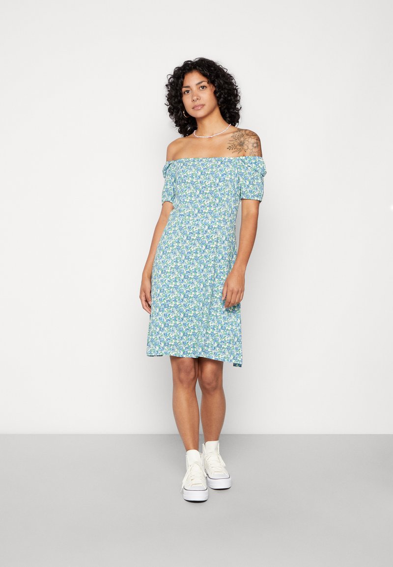 Vestido azul claro de flores, com alças à descoberta dos ombros e mangas bufantes curtas, até ao joelho, feito de um tecido suave, combinado com ténis brancos de cano alto.