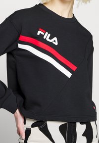 Personne portant un sweat-shirt noir Fila avec des rayures diagonales rouges et blanches et un pantalon à motifs noir et blanc, debout avec la main sur la hanche.