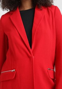 Femme portant un blazer rouge vif avec des revers, deux poches zippées, sur un top noir, coupé pour montrer le haut du torse et les épaules.