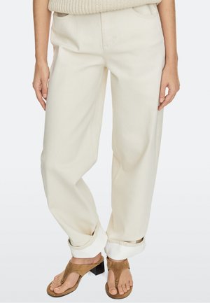 Personne portant un pantalon crème à jambes larges retroussé aux ourlets avec des sandales à talons en daim marron et un pull en maille beige.