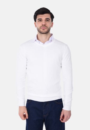 Maglione bianco a collo aV in tessuto lavorato a maglia, con polsini e orlo a coste, indossato sopra una camicia azzurra con colletto, abbinato a jeans scuri.