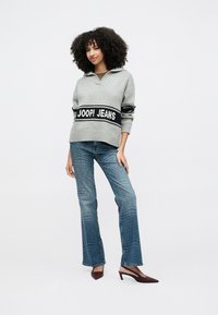 Frau mit lockigem Haar, die einen grauen Pullover mit der Aufschrift "JOOP! JEANS", blaue Jeans mit wiederholtem Logo und burgunderrote Absätze trägt, vor weißem Hintergrund stehend.