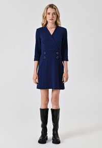 Navyblaues Wickelkleid mit Dreiviertelärmeln, doppelten Knopfdetails und ausgestelltem Schnitt, kombiniert mit knielangen schwarzen Stiefeln.