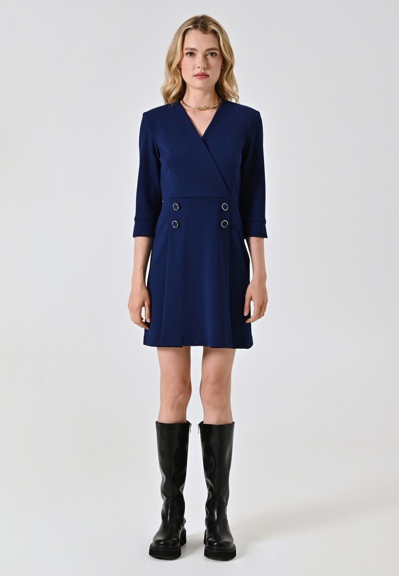 Navyblaues Wickelkleid mit Dreiviertelärmeln, doppelten Knopfdetails und ausgestelltem Schnitt, kombiniert mit knielangen schwarzen Stiefeln.