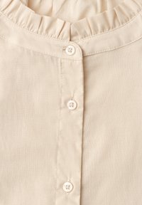Blouse beige en gros plan montrant trois petits boutons ronds et un détail de col volanté froncé.