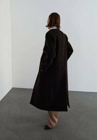 Abrigo largo marrón oscuro con mangas oversized, forma estructurada y aberturas laterales, combinado con pantalones beige y zapatos negros de punta abierta.
