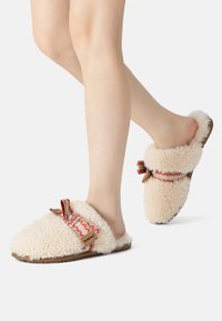 Fuzzy beige slippers made of soft shearling, featuring decorative red and white patterned straps and a brown leather accent.