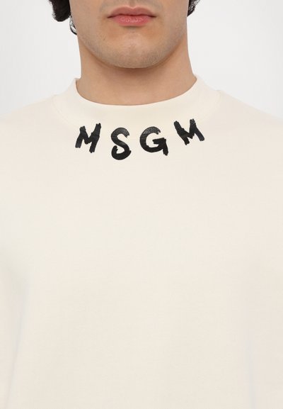 Sweatshirt crème avec encolure ronde. Le devant présente les lettres noires brodées "MSG" dans une police épaisse et inégale. Tissu doux et texturé.