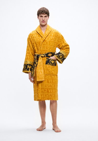 Rumeno kopalno haljo iz frotirja z izbočenim vzorcem logotipa "VERSACE", črnim satenastim pasom in kontrastnimi črnimi in zlatimi poudarki na rokavih.