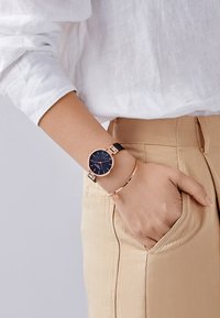 Montre noire avec un cadran bleu rond et des accents en or rose, portée avec un bracelet en or rose, sur une main dans une poche de pantalon beigne.