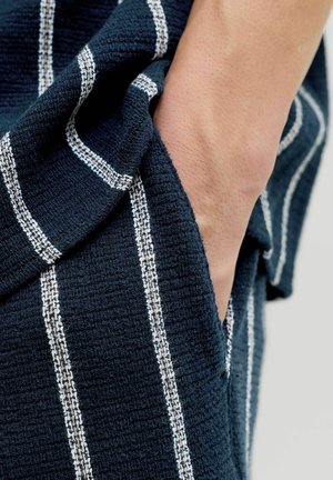 Käsi tumesinise tekstuuriga kanga taskus, millel on valged vertikaalsed triibud, näidates osa käsivarrest ja randmest.