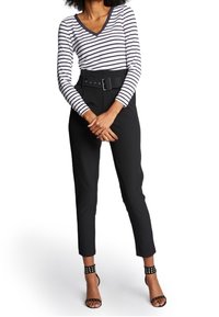 Kvinna klädd i svarta högmidjade byxor med bälte, svarta spikade ankelrem-sandaler och en vit långärmad skjorta med svarta horisontella ränder.