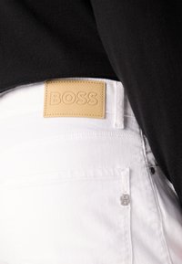 Baltas denims ar vieglu brūnu ādas uzlīmi, uz kuras ir iegravēts "BOSS". Iespējas ietver sudrabu pogu un detalizētu šuvumu.