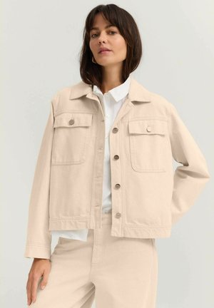 Beige Jeansjacke mit zwei Brusttaschen, silbernen Knöpfen und einem klassischen Kragen, über einem weißen Hemd getragen. Glatte Textur.