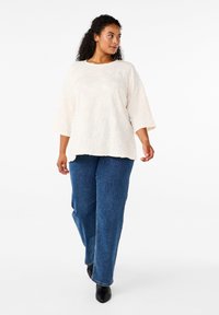 Top bianco texturizzato con maniche a 3/4 abbinato a jeans a gamba larga blu e calzature nere, su un modello in piedi contro uno sfondo bianco.