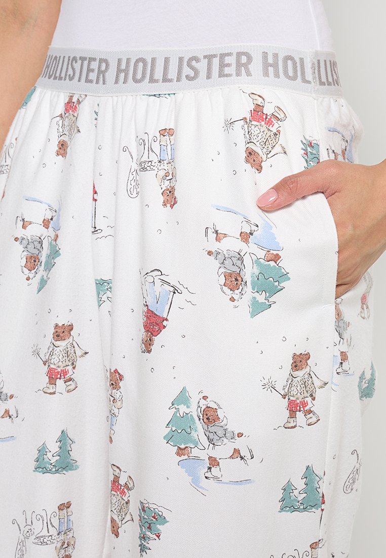 Weiße Schlafanzughosen mit einem grauen "Hollister"-Bund, der eine wiederholte Illustration von Cartoon-Bären in winterlichen Szenen zeigt.