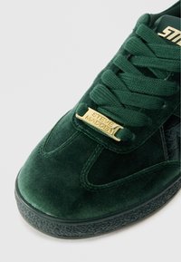 Zielone sneakersy z weluru z teksturowanym czarnym boku, płaskimi sznurówkami i złotą metalową płytką z logo na języku. Gumowa podeszwa z wzorem antypoślizgowym.