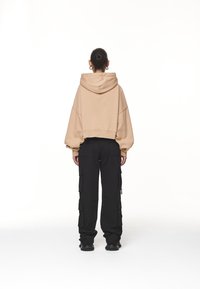 Person med knut i håret iförd beige hoodie och svarta cargo byxor, stående vänd bort från kameran mot en vit bakgrund.