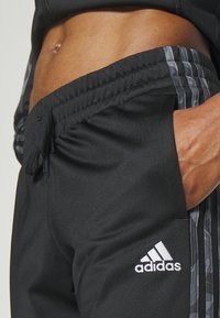 Čierne športové nohavice vyrobené z hladkej tkaniny, s elastickým pásom, bočnými vreckami a bielym logom Adidas so šedými pruhmi.