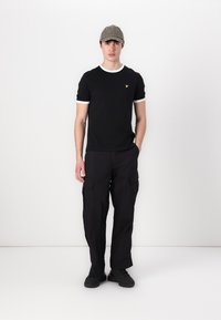 Czarny t-shirt z białymi akcentami kołnierza i małym żółtym logo, zestawiony z luźnymi czarnymi spodniami cargo i czarnymi sneakersami.