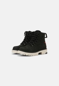 Botas de tobillo de ante negro con suela de goma beige, diseño con cordones y tirador trasero, que cuentan con ojales metálicos y acentos texturizados.