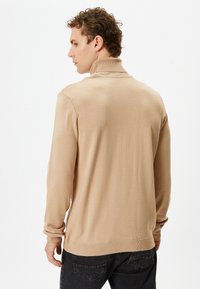 Beige gebreide trui met een hoge kraag en lange mouwen. Heeft een gladde textuur en een geribde zoom, gecombineerd met donkere jeans.