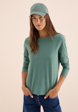 T shirt à manches longues, col arrondi, vert en tissu doux, assorti à une casquette verte. Porté avec un jean en denim bleu.