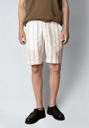 Man die beige en wit gestreepte korte broek, een bruin overhemd en zwarte veterschoenen draagt, staand tegen een effen lichtgrijze achtergrond.