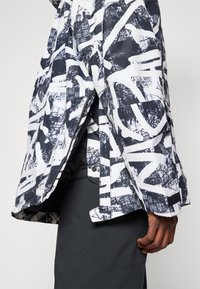 Μπουφάν με επένδυση σε μαύρη και λευκή αφηρημένη σχέδιο, διαθέτει κλείσιμο με φερμουάρ και πλαινά σκισίματα, κατασκευασμένο από ελαφρύ, αδιάβροχο υλικό.