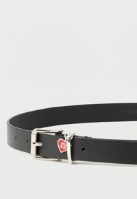 Must nahast vöö hõbedase pandlaga, millel on südamekujuline ripats tekstiga "HUGO". Siidise tekstuuriga ning ühtlaselt paigutatud augud reguleerimiseks.