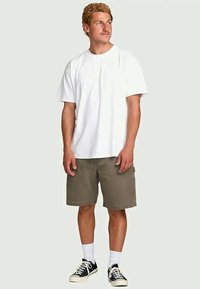 Young man standing, wearing a white t-shirt, olive green knee-length shorts, white socks, and black sneakers, looking to his left.
