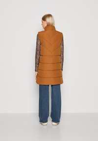 Brun puffer vest med høy krage, med vertikal søm og lommer, båret over en mønstrete langermet topp med vide denimbukser.