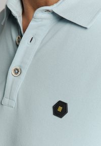 Lichtblauw polo-shirt van een gestructureerde stof, met een zwarte hexagonale patch met gele details en twee knopen op de kraag.