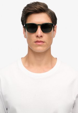 Joven con cabello corto castaño que lleva gafas de sol con montura verde oscuro y una camiseta blanca lisa de cuello redondo contra un fondo blanco.