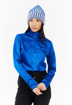 Blauwe, langlopende, rits-top gemaakt van glanzend materiaal, gecombineerd met een gebreide blauw-witte gestreepte beanie. Zwarte broek met voorkeurszakken.