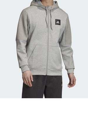 Hombre vistiendo una sudadera con capucha Adidas de color gris claro con cremallera, capucha y bolsillo estilo canguro, combinado con pantalones gris oscuro.