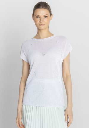 Kobieta z jasnobrązowymi włosami związanymi do tyłu, ubrana w biały, krótki sweter z subtelnymi diamentowymi wzorami oraz jasną plisowaną spódnicę.