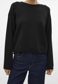 Haut noir à manches longues en tissu texturé avec un col rond et un ourlet légèrement raccourci, associé à un jean bleu foncé.