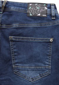 Jeans de denim en un tono azul oscuro, con costuras amarillas, un bolsillo trasero con forma puntiaguda y una etiqueta texturizada con acentos geométricos coloridos.