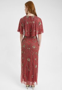 Lungo vestito bordeaux con una mantellina trasparente, decorato con ricami floreali dorati e motivi a foglia. Include una chiusura con zip sul retro.