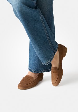 Person wearing blue jeans and brown suede loafers, one foot crossed over the other against a white background.