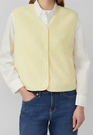 Personne portant un gilet polaire jaune clair boutonné par-dessus une chemise blanche à manches longues et un jean bleu, main dans la poche sur un fond gris.