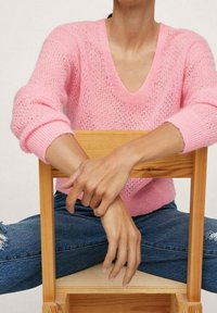 Maglione rosa chiaro a maglia con scollo a V e polsini a costine, abbinato a jeans blu strappati. La modella è seduta su una sedia di legno.