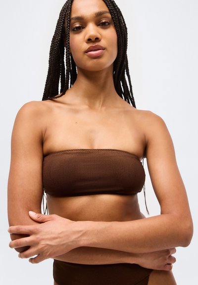 Kobieta z długimi splecionymi warkoczami, ubrana w brązowy top bez ramiączek, stojąca z założonymi na siebie rękami na tle jednolitego jasnoszarego tła.