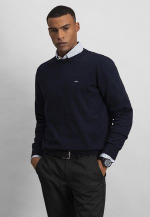 Mann trägt einen marineblauen Pullover über einem weißen, gemusterten Hemd, dunkelgraue Hose, schwarzen Gürtel und schwarze Armbanduhr, steht mit den Händen in den Taschen.