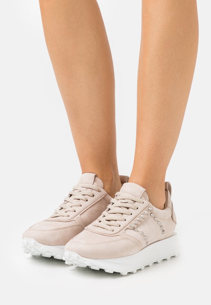 Ljusbeige sneakers i mocka, med dekorativa nitar på sidan, vit sula och traditionell snörning.