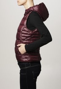 Bordeauxrote Steppweste mit Kapuze, die ein gesteckte Muster und ärmellosen Schnitt aufweist. Weiche Textur mit Seitentaschen und einer taillierten Silhouette.