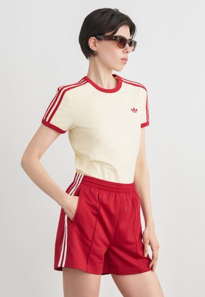 Kobieta w kremowo-czerwonej koszulce Adidas oraz pasujących czerwonych szortach i okularach przeciwsłonecznych, stojąca z jedną ręką w kieszeni, zwrócona w prawo.