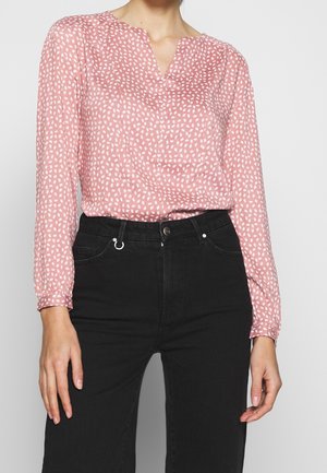 Femme portant un chemisier rose à manches longues à pois blancs, rentré dans un pantalon noir taille haute, debout avec les mains détendues le long du corps.