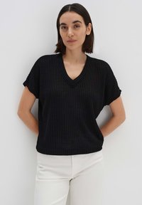 Schwarzes, geripptes kurzärmliges Oberteil mit einem V-Ausschnitt. Der Stoff hat eine strukturierte Oberfläche. Getragen mit hellen Hosen, die Hände auf den Hüften.